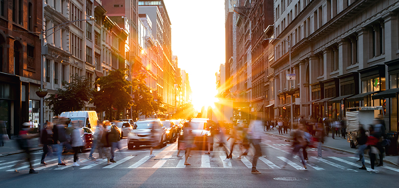 Crowds of busy people walking through the intersection of 5th Avenue and 23rd Street in Manhattan, New York City with bright sunset background