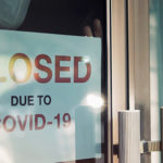 Business office or store shop is closed/bankrupt business due to the effect of novel Coronavirus (COVID-19) pandemic. Unidentified person wearing mask hanging closed sign in background on front door.