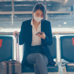 business lady traveller wear suit sitting with suitcase and use smart phone chat message in bench wait for flight at airport. Business travel commuter in covid pandemic, Business travel concept.