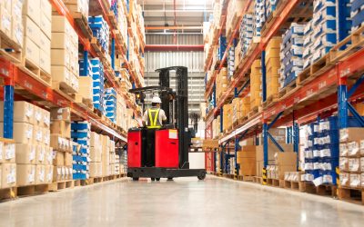 Worker in forklift-truck loading packed goods in huge distribution warehouse with high shelves.