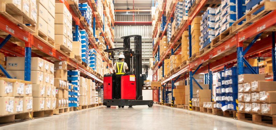 Worker in forklift-truck loading packed goods in huge distribution warehouse with high shelves.