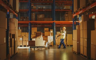 Worker Moves Cardboard Boxes using Hand Pallet Truck, Walking between Rows of Shelves with Goods in Retail Warehouse. People Work in Product Distribution Logistics Center. Side View Shot