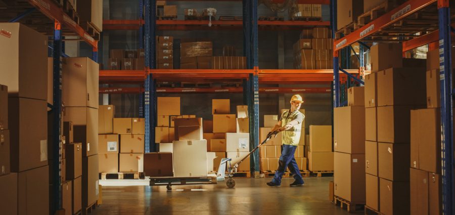 Worker Moves Cardboard Boxes using Hand Pallet Truck, Walking between Rows of Shelves with Goods in Retail Warehouse. People Work in Product Distribution Logistics Center. Side View Shot