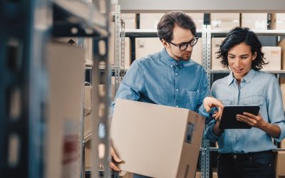 Female Inventory Manager Shows Digital Tablet Information to a Worker Holding Cardboard Box, They Talk and Do Work. In the Background Stock of Parcels with Products Ready for Shipment.