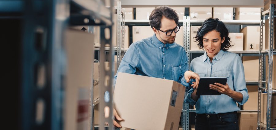 Female Inventory Manager Shows Digital Tablet Information to a Worker Holding Cardboard Box, They Talk and Do Work. In the Background Stock of Parcels with Products Ready for Shipment.