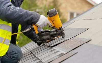 Unrecognizable roofer worker in special protective work wear and gloves, using air or pneumatic nail gun and installing asphalt or bitumen shingle on top of the new roof under construction residential building