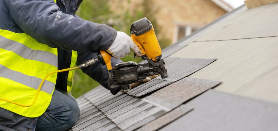 Unrecognizable roofer worker in special protective work wear and gloves, using air or pneumatic nail gun and installing asphalt or bitumen shingle on top of the new roof under construction residential building