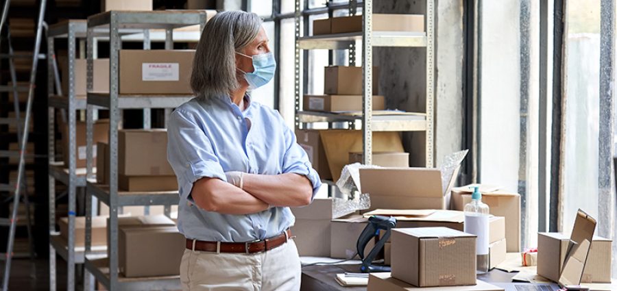 Older mature female online store small business owner, manager, stock worker, entrepreneur wearing face mask and gloves standing with arms crossed at workplace in warehouse looking through window.