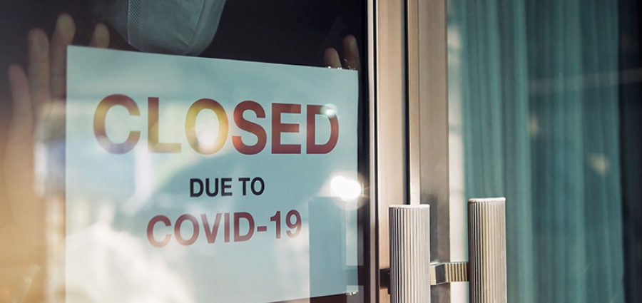 Business office or store shop is closed/bankrupt business due to the effect of novel Coronavirus (COVID-19) pandemic. Unidentified person wearing mask hanging closed sign in background on front door.