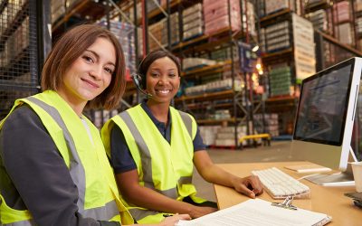 Two female colleagues in a warehouse office look to camera