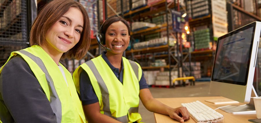 Two female colleagues in a warehouse office look to camera